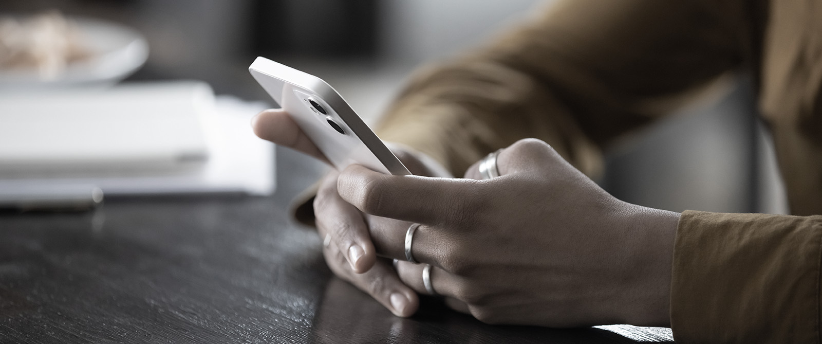 The image shows a person holding a smartphone with both hands, focusing on the screen while seated at a table, with a blurred background suggesting an indoor setting.