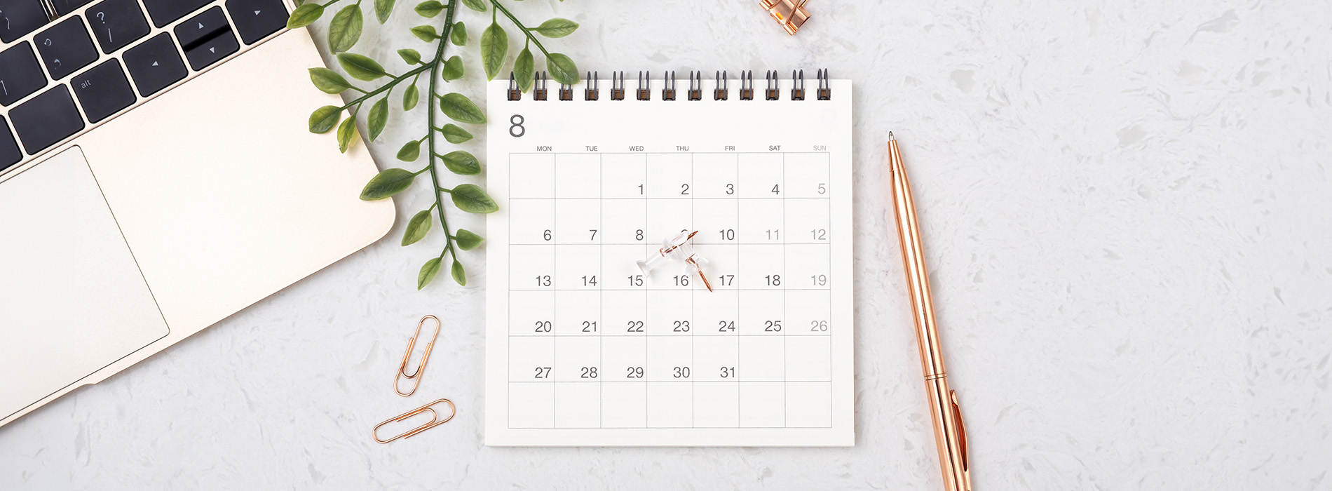 The image displays a calendar with a pen on top, placed on a surface with a laptop and a plant in the background.