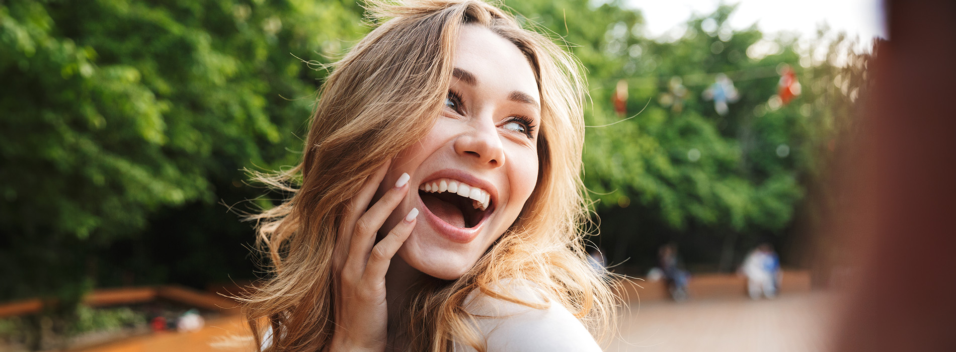 Woman with blonde hair smiling at camera, wearing a white top and sunglasses, outdoors during daylight.