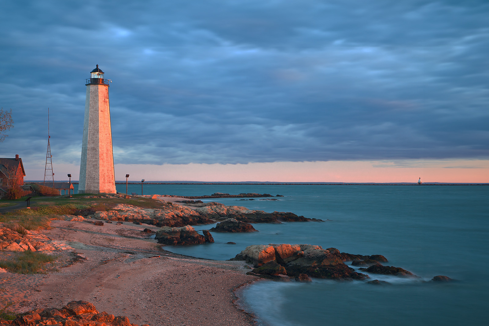 The image captures a serene coastal scene at sunset, featuring a lighthouse on a rocky shore with calm waters and a clear sky.